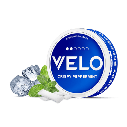 Un envase redondo azul y blanco de VELO Crispy Peppermint se muestra con hojas de menta, cubitos de hielo y bolsitas de nicotina blanca de origen vegetal dispuestas delante sobre un fondo blanco.