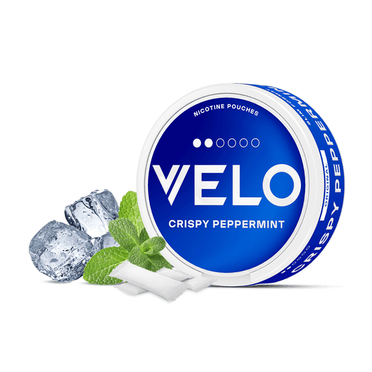 Un envase redondo azul y blanco de VELO Crispy Peppermint se muestra con hojas de menta, cubitos de hielo y bolsitas de nicotina blanca de origen vegetal dispuestas delante sobre un fondo blanco.